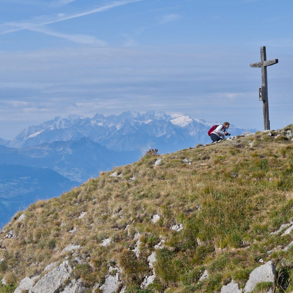A hiker pauses at a mountain peak marked by a cross, overlooking stunning Alpine views.